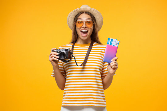 Happy excited young female tourist holding passport with tickets and camera, isolated on yellow background