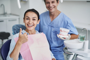 Charming lady doing thumbs up sign while dentist standing behind her