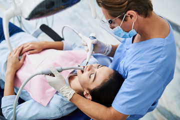 Competent dentist in protective mask doing teeth cleaning for young lady