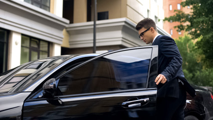Handsome office worker getting in new luxury car near business center, promotion