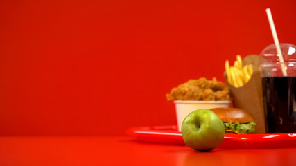 Green apple lying on table, fast food on background, choosing healthy nutrition