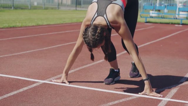 Slow motion tilt up of young female sprinter being in starting position prepared for run and beginning her race on track at outdoor stadium