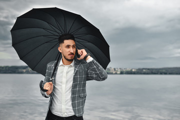 A good deal! Bearded businessman speaks by phone and laughs.View of a Young attractive business man holding the umbrella using smartphone. Smiling formal dressed businessman talking on cell phone with