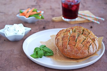 Party set on a brown concrete background: baked party bread with herbs and olive oil, a dip of cream cheese with vegetables, sliced raw carrots and green sweet pepper, grape juice with ice, straw