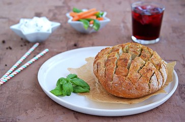 Party set on a brown concrete background: baked party bread with herbs and olive oil, a dip of cream cheese with vegetables, sliced raw carrots and green sweet pepper, grape juice with ice, straw