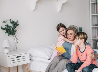 Cropped photo of gray-haired woman reading book for her grandchildren in house