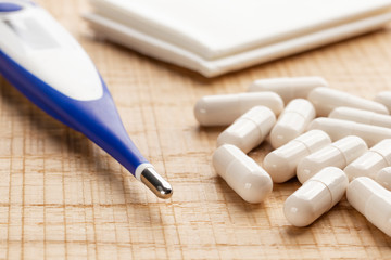 Electronic thermometer and pills with paper handkerchiefs on wooden background.