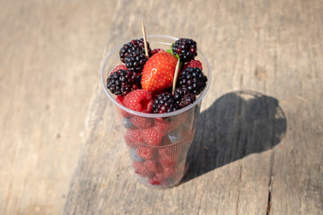 Plastic cup with toothpicks and mixed berries -  blackberries, raspberries, strawberry on wooden table