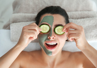 Pretty woman with clay mask on her face holding cucumber slices in spa salon, above view