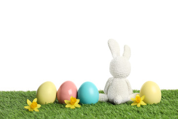 Cute Easter bunny toy and dyed eggs on green grass against white background, space for text