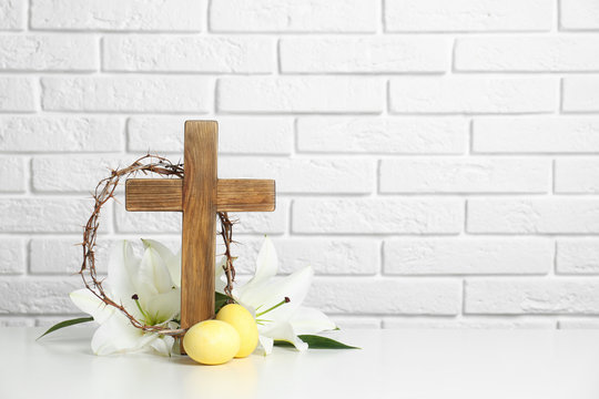 Wooden cross, crown of thorns, Easter eggs and blossom lilies on table against brick wall, space for text
