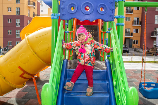 The little girl, warmly dressed, in a hat and jacket plays on the playground with slides and swings in the courtyard of residential buildings.