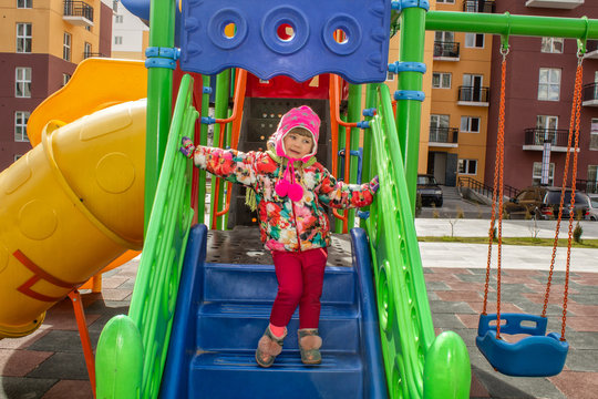 The little girl, warmly dressed, in a hat and jacket plays on the playground with slides and swings in the courtyard of residential buildings.