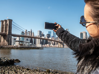 Young Woman tourist by the river in Dumbo taking pictures of Brooklyn Bridge and Cityscape of New...