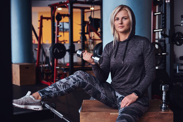 Fototapeta na wymiar Beautiful sportive girl with blonde hair wearing hoodie sitting on a wooden box and holding a dumbbell in the gym