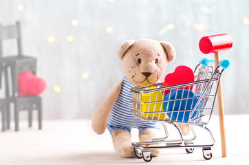 Teddy bear looking at shopping cart with children's wooden toys and hammer. Fairy lights background