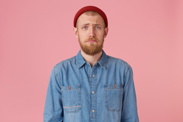 Portrait of blue-eyed young guy in red hat with red beard looks sad, upset, frustrated, displeased...