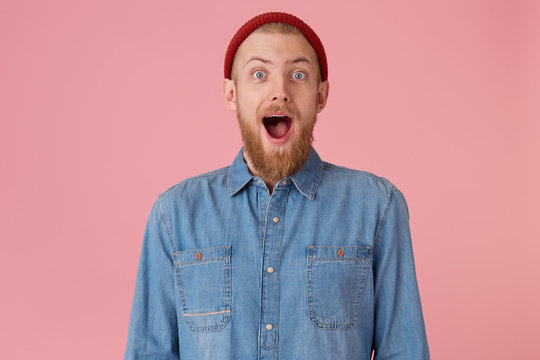Overflowing with positive emotions glad guy in red hat with red thick beard openes mouth widely in excitement, dropped jaw, isolated on pink wall