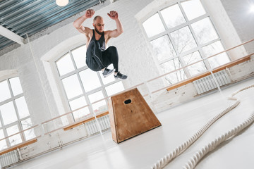 Dynamic shot of fitness male athlete jumping through at square box in crossfit gym. Strong man doing jump on wooden squat box at light hall