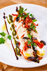 Caprese salad with tomatoes, mozzarella cheese, pesto sauce and black olives on white plate. Close up