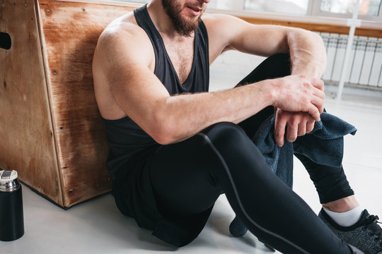 Tired Strong Man Sitting On Box At Gym. Perspiring Fit Sportsman Resting With Towel After Hard Crossfit Workout In Light Hall