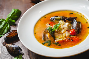 Soup with mussels and vegetables in white plate on wooden table. Close up