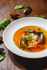 Soup with mussels and vegetables in white plate on wooden table. Close up