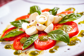 Traditional Italian caprese salad with sliced tomatoes, mozzarella cheese, basil and pesto sauce on white plate. Close up