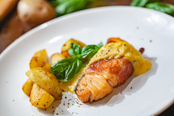 Grilled salmon fillet wrapped in bacon and potato wedges on white plate. Close up