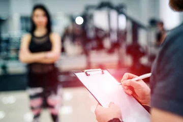 Tuinposter Trainer and client discussing her progress at the gym. Gym instructor writing on clipboard © Vadim