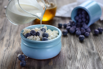 Fresh rustic cottage cheese in a blue cup with blueberries and cream on a wooden table next to scattered blueberries and a jar of honey