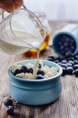 Fresh rustic cottage cheese in a blue cup with blueberries and cream on a wooden table next to scattered blueberries and a jar of honey