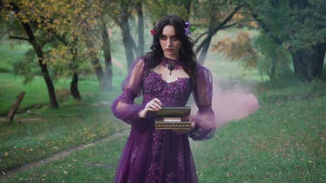 brunette girl with dark make-up poses for camera in amazing chic seductive purple dress with open neckline, lady with flowers in hair holds mysterious casket with pink smoke. Punishment for pandora