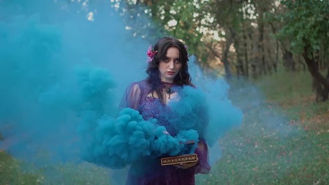 portrait fantasy woman greek goddess Pandora holding magic box in hands, filming with colored blue smoke bombs. Purple dress crown wreath. Gothic evil queen girl. Mystic summer nature forest tree