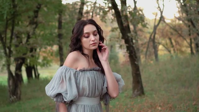 brunette girl in free gray dress with open bare shoulders in forest and posing for camera photo shoot, natural nude makeup and light hair styling, witch drank magic potion to became young beauty