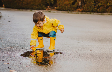 Cute playful little baby boy in bright yellow raincoat and rubber boots playing with rubber ducks in small puddle at rainy spring day on wet street road. Seasonal weather walking concept