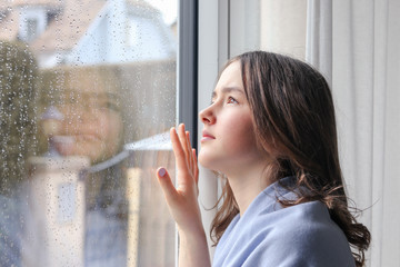 Beautiful melancholic teenager girl in light blue shawl  looking outside through raindrops on wet window at sky at spring rainy day. Seasonal weather concept.