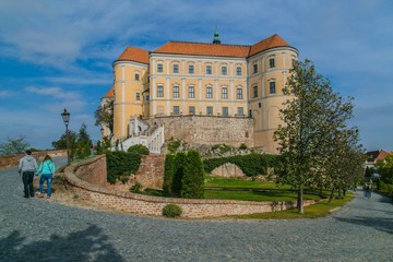 Mikulov, Czech Republic / South Moravia - October 15 2016: Mikulov castle with yellow and white facade and red roof standing on a rock, green vegetation in garden, tourists walking on pavement
