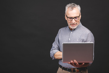 Senior aged bearded old man in eyeglasses holding laptop computer and smiling isolated against black background.