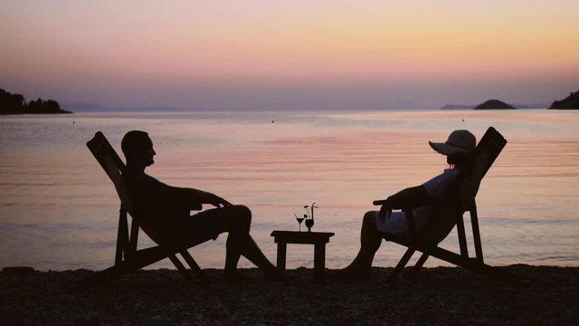 Romantic couple silhouettes sit on chair and relax at sunset, cocktail glasses