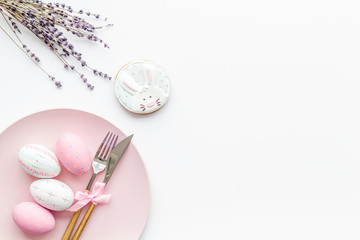 Easter dining table in pastel colors. Plate, cutlery, painted eggs, gingerbread and lavender branches on white background top view copy space