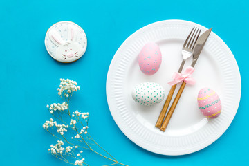 Easter dining table in pastel colors. Plate, cutlery, painted eggs, gingerbread and dry flower branch on blue background top view