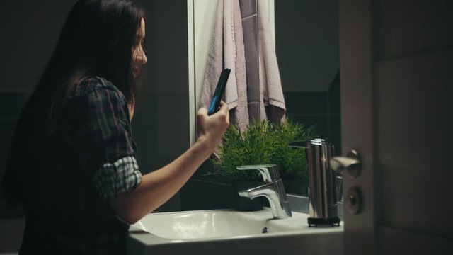 Young woman taking selfie in front of mirror in the bathroom at night, camera on slider