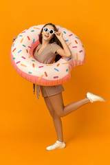 Vertical full length picture of stylish teenage girl in summer dress, sunglasses and sneakers posing with inflatable pink circle, going to have swim in swimming pool during vacations in resort area
