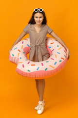 Vacation, summertime and water activity concept. Vertical image of beautiful cheerful young lady wearing shades and striped dress posing isolated with pink inflatable swimming circle around her waist