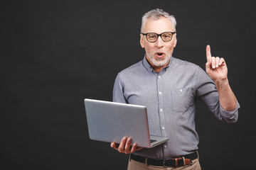 Great idea! Happy senior aged man pointing up at copy space while holding laptop isolated over black background.