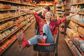 Young couple in grocery store. Woman sit in trolley and have fun. Man stand behind and point on shelf with products. Happy couple having fun.