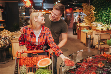 Young couple in grocery store. She get plate with berries and look at man. Guy Stand behind her and...