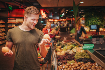 Young couple in grocery store. Man stand in front and hold kiwi in hands. Woman get pineapple behind. They look happy.