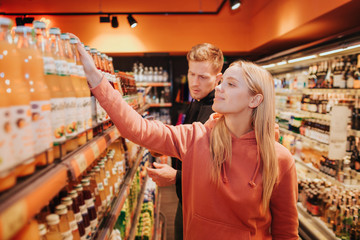 Young couple in grocery store. She pick up juice bottles and touch them with hand. Serious concentrated man stand behind and look down. Grocery shopping.